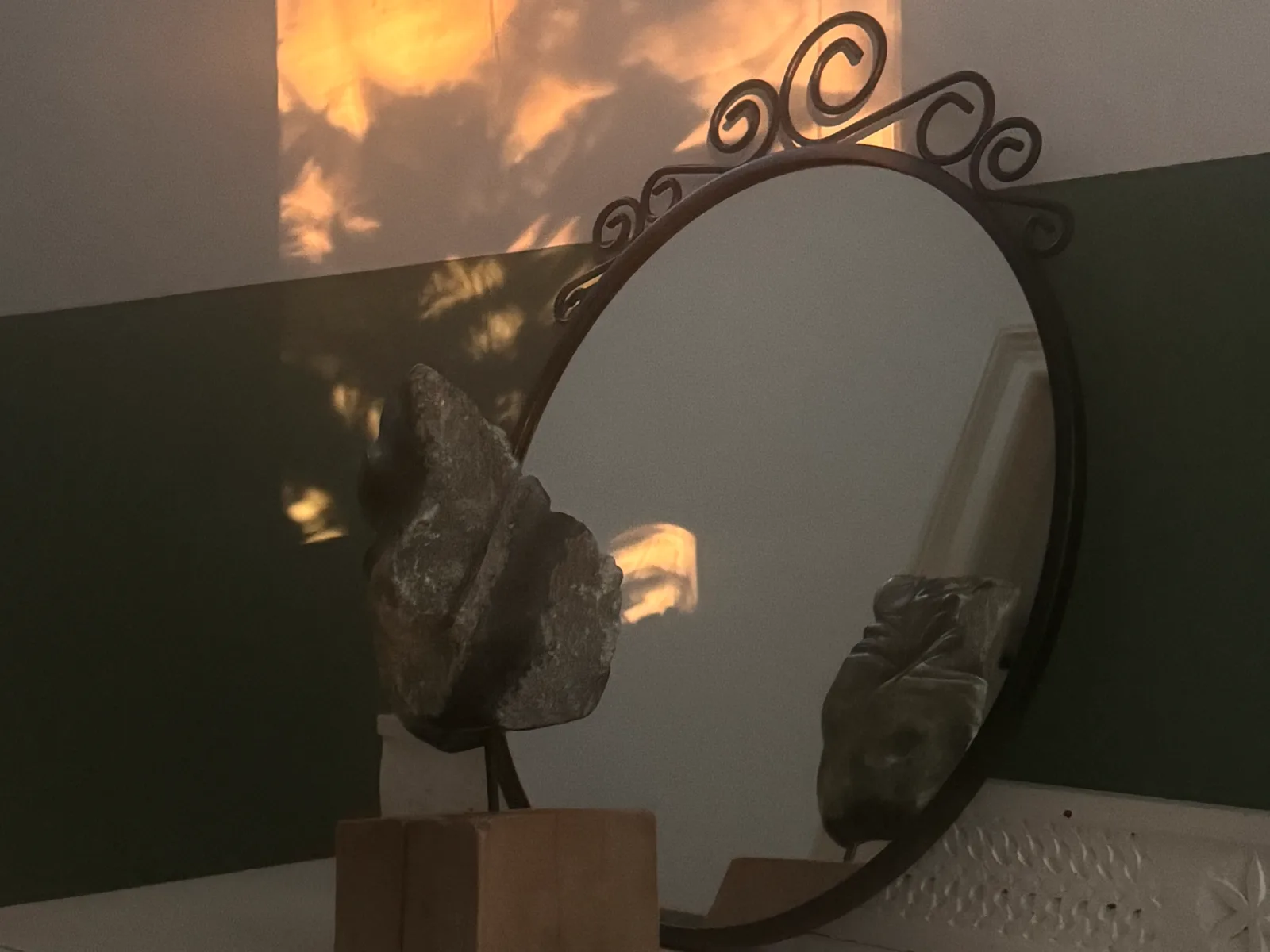 Beeld in speksteen op een houten voet voor een ronde spiegel, met avondlicht op de muur